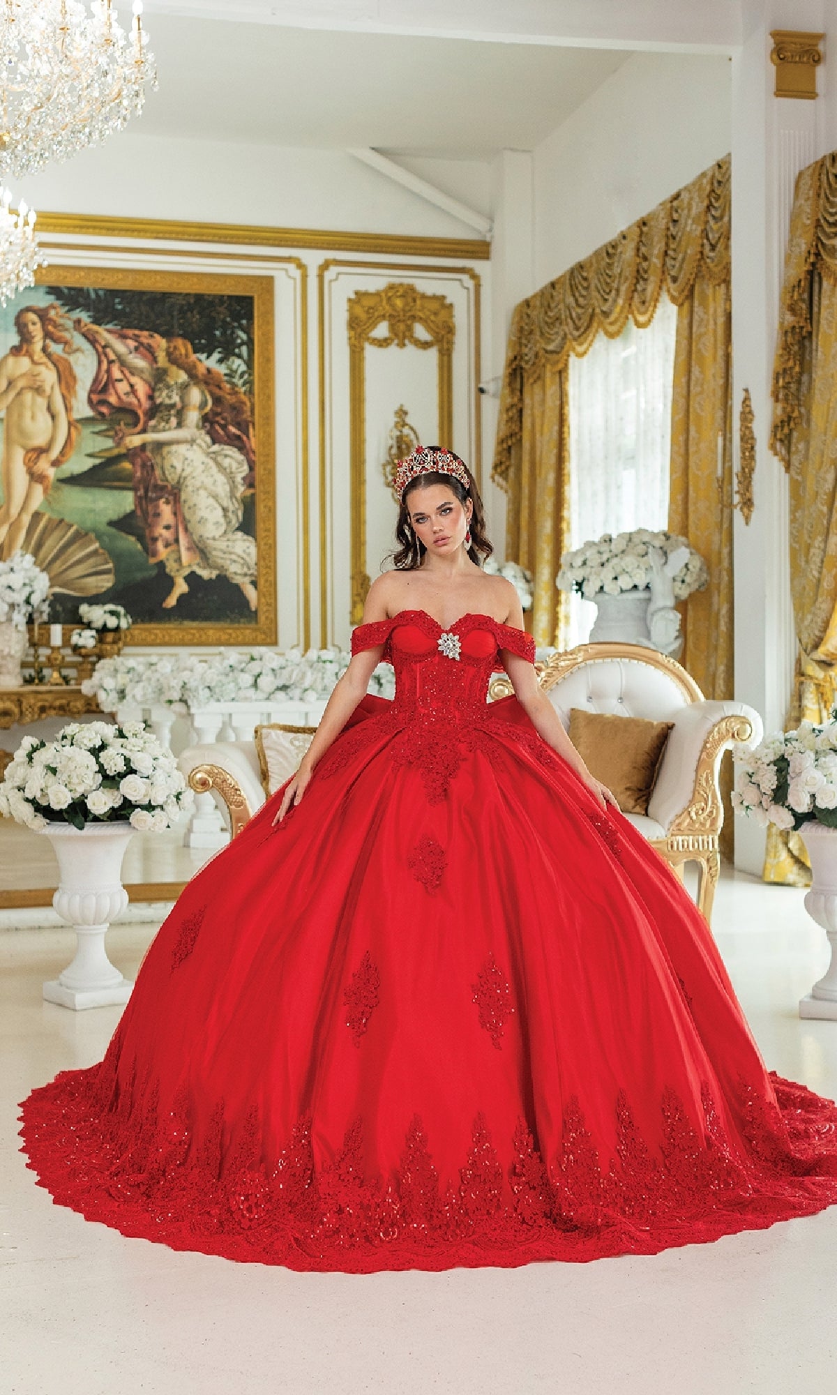 A woman in the Quinceanera Dress 1913 by Dancing Queen, a red off-the-shoulder gown with a corset bodice and tiara, stands before a gold-trimmed sofa, surrounded by white flowers and a large classical painting in the background.
